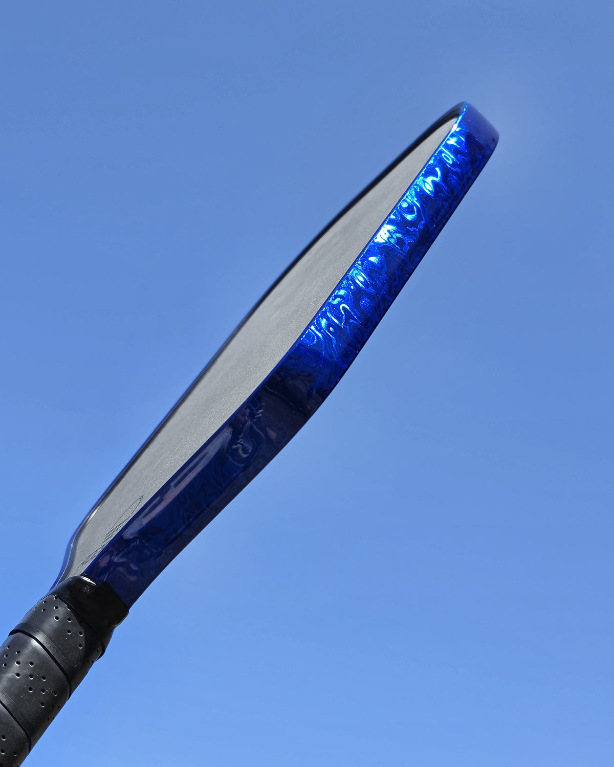 A close-up shot of a pickleball paddle showing its vibrant Blue Damascus edge guard tape against a clear blue sky background.