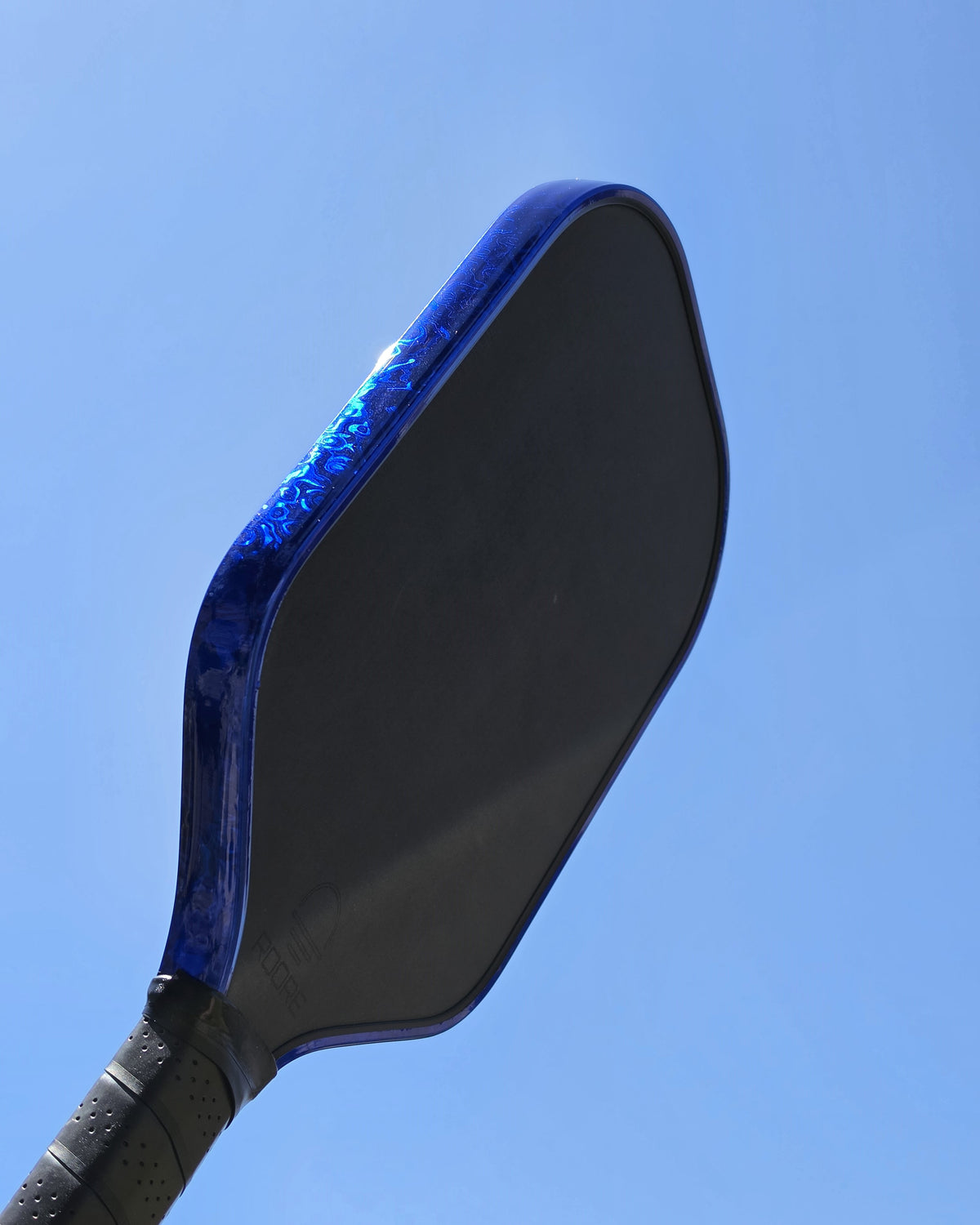 A close-up shot of a pickleball paddle against a clear blue sky. The paddle features a vibrant Blue Damascus finish protective edge tape.