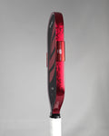 A close-up side profile of a pickleball paddle showing a Red Damascus edge tape against a gray background.