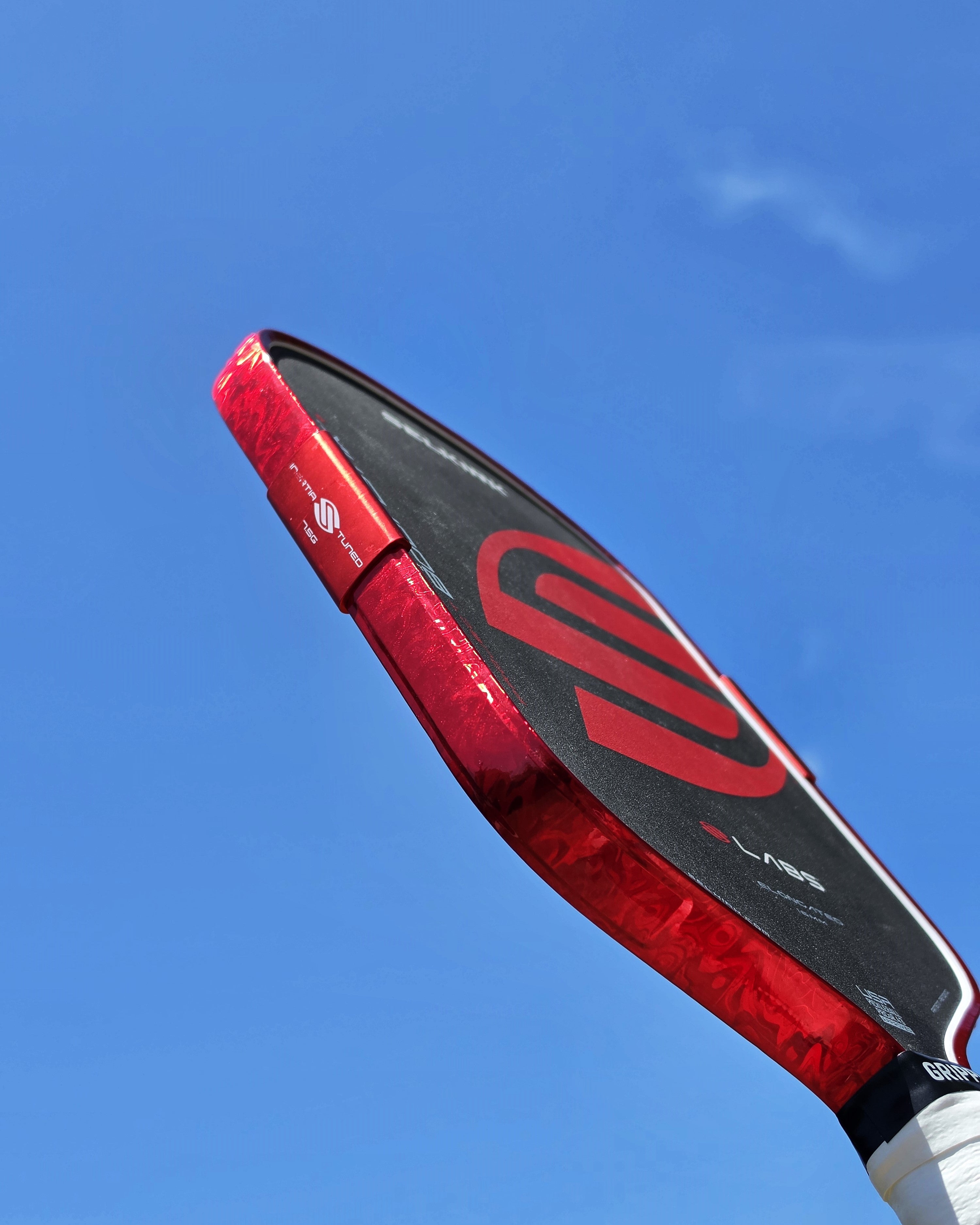 A close-up, low-angle shot of a Boomstik pickleball paddle with Red Damascus edge tape installed shown against a clear blue sky background.