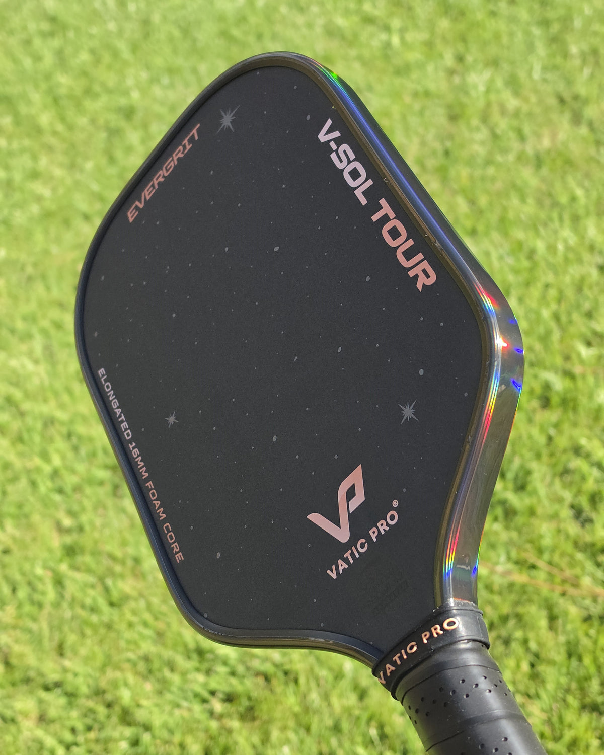Close-up of a VATIC PRO V-Sol Tour pickleball paddle, showing the Smoke Hologram Ultimate Edge Tape outdoors against a green grass background.