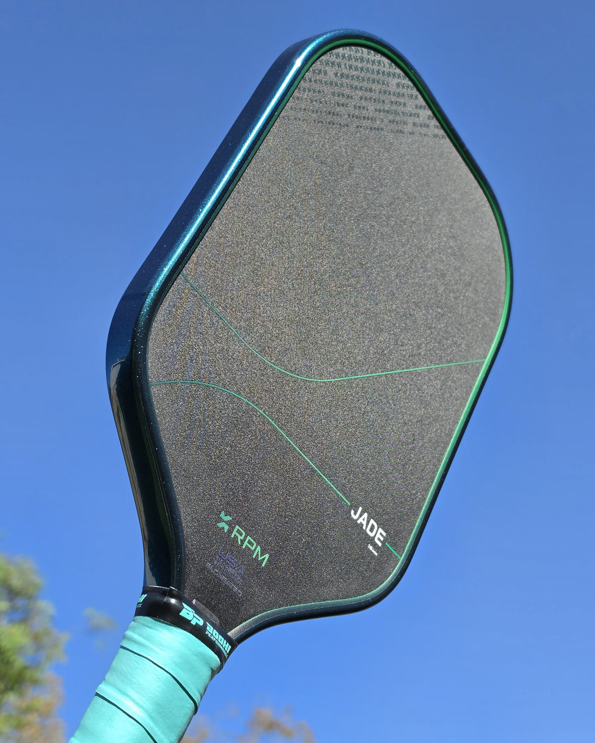 RPM Jade pickleball paddle with teal jade metallic edge tape over a sky background