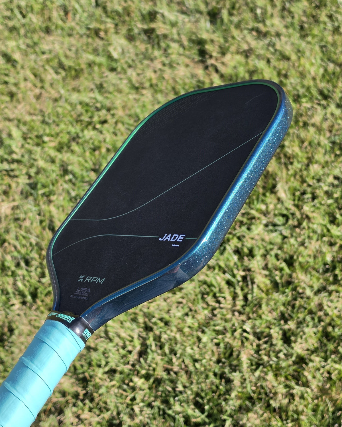 RPM Jade pickleball paddle with teal jade metallic edge tape over a grass background.